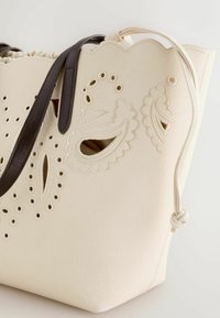 Sac fourre-tout crème avec bords festonnés, broderie florale en découpe et anses en cuir marron foncé contrastant.