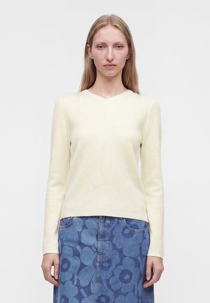 Vrouw met steil blond haar draagt een crème kleurige top met lange mouwen en een blauwe rok met grote bloemenpatronen, staand tegen een effen achtergrond.