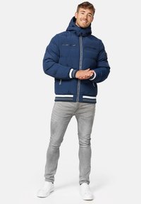 Mørkeblå puffjakke med hætte, to brystlommer med lynlås, ribbede manchetter og nederste kant med hvide striber. Båret over lysegrå jeans og hvide sko.