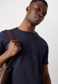 Navyblaue Baumwoll-T-Shirt mit Rundhalsausschnitt und ausgefransten Kanten, getragen von einer Person, die eine braune Umhängetasche mit einem Riemen hält.