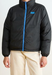 Svart puffjacka med hög krage, med en blå dragkedja och logo. Elastiska ärmslut och en något kortad design. Slät textur.