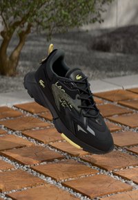 Black and yellow Lacoste sneaker with laces, floating above wooden tiles and gravel outdoors near a small tree.