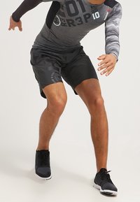 Reebok sportovní oblečení sestávající z těsně padnoucího šedého dlouhého rukávu s černými a maskáčovými akcenty, spárovaného s volnými černými šortkami a sportovními černými botami.