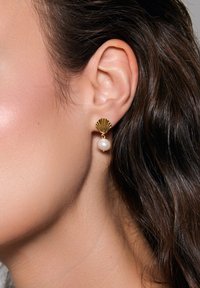 Złote kolczyki w kształcie muszelek z białą perełką, podkreślające gładkie tekstury i kontrastowy design.