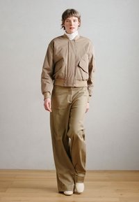 Tan bomber jakna z dvema sprednjima žepoma, kombinirana z ohlapnimi hlačami v isti barvi. Model nosi belo majico z visokim ovratnikom in bež čevlje.