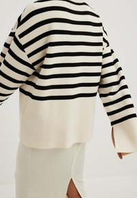 Pull rayé noir et crème avec une coupe ample, une texture côtelée et des manches larges, complété par un détail d’ourlet fendu en bas.