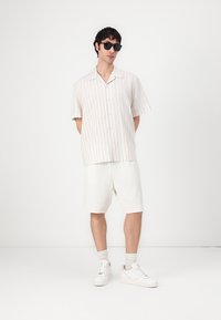 Κοντομάνικο πουκάμισο με κουμπιά σε ανοιχτό ριγέ σχέδιο σε μπεζ χρώμα, συνδυασμένο με λευκά σορτς και λευκά αθλητικά παπούτσια, φορεμένο με σκούρα γυαλιά ηλίου.