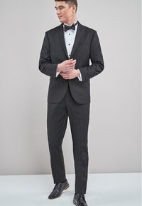 Schwarzer Smoking mit schimmerndem Revers, weißes Hemd und schwarze Fliege. Enganliegende Hosen und glänzende schwarze Lackschuhe mit braunen Akzenten.