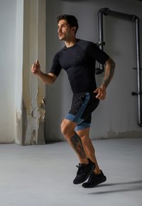 Juoda prigludusi sportinė marškinėlė ir juodi šortai su mėlynais akcentais. Bėgikas juda, demonstruodamas tatuiruotes ir juodus sportinius batus ant lygaus grindų paviršiaus.