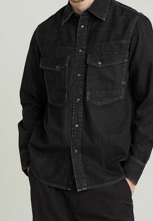 Mann trägt ein schwarzes, button-up Denim-Hemd mit zwei Brustklappentaschen und steht mit den Händen in den Taschen vor einem neutralen Hintergrund.