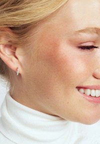 Leende kvinna med slutna ögon, ljus hy, blont hår, iförd små silverfärgade hoopörhängen och en vit ribbad polotröja.