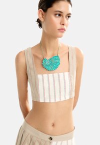 Beżowy crop top w czerwone paski, zestawiony z beżową spódnicą. Stylizacja uzupełnia turkusowa, teksturowana naszyjnik w kształcie muszli.