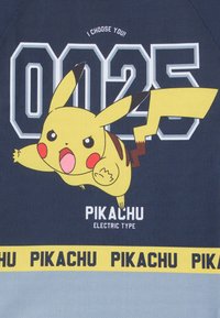 Tamsiai mėlynas grafikos marškinėlis, vaizduojantis Pikachu geltonoje spalvoje, su užrašu "0025" ir "AŠ TAVE PASIRINKU!". Geltona juosta apačioje skelbia "PIKACHU."