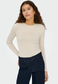 Langermet beige topp laget av mykt stoff, med en bølget utringning, kombinert med mørkeblå jeans.