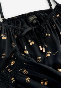 Tissu noir avec imprimé cerisiers dorés, doté d'une taille élastique et de bretelles réglables, avec une étiquette logo et des accessoires métalliques.