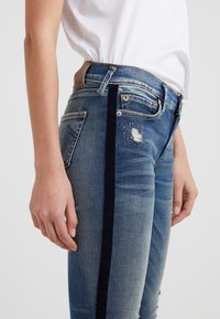 Person som bär blå denimjeans med en svart sidremsa, sliten patch med metallstift, och en vit inskjuten skjorta.