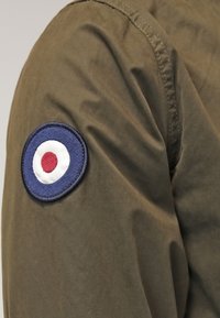 Olivgrön jacka med en rund broderad patch på ärmen med en blå kant och en central röd prick inom en vit cirkel.