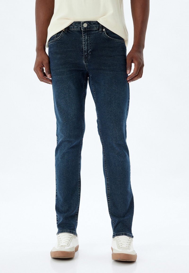 Jeans de mezclilla azul oscuro con un corte recto, con estilo de cinco bolsillos y cierre de botón, combinados con zapatillas blancas.