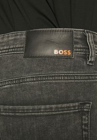 Närbild av mörkgrå denimjeans med en svart läderlapp märkt "BOSS" vid midjan och en svart skjorta instoppad.