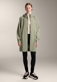 Jachetă impermeabilă verde deschis, cu glugă, fermoar pe toată lungimea și buzunare laterale, purtată cu colanți negri și adidași. Textură netedă, design casual.