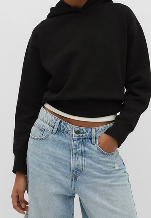 Person trägt einen schwarzen kurzen Hoodie über einem weißen Unterhemd und hellblaue High-Waist-Jeans mit einer Hand in der Tasche.