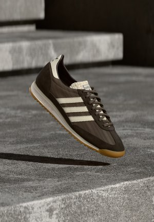 Brauner Sportschuh mit hellbeigen Streifen, Veloursleder- und Mesh-Material, strukturierter Oberseite, Schnürdesign und Laufsohle aus Gummibelag.