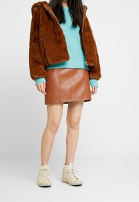 Veste à capuche en fausse fourrure marron, pull en maille vert menthe, mini-jupe en cuir marron, baskets montantes beiges. Textures et couleurs nettes et modernes.