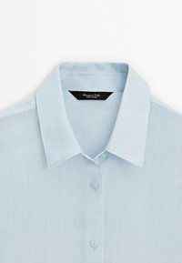 Ljusblå linneskjorta med en strukturerad krage, central knappslå och en etikett på kragen med varumärkesinformation.