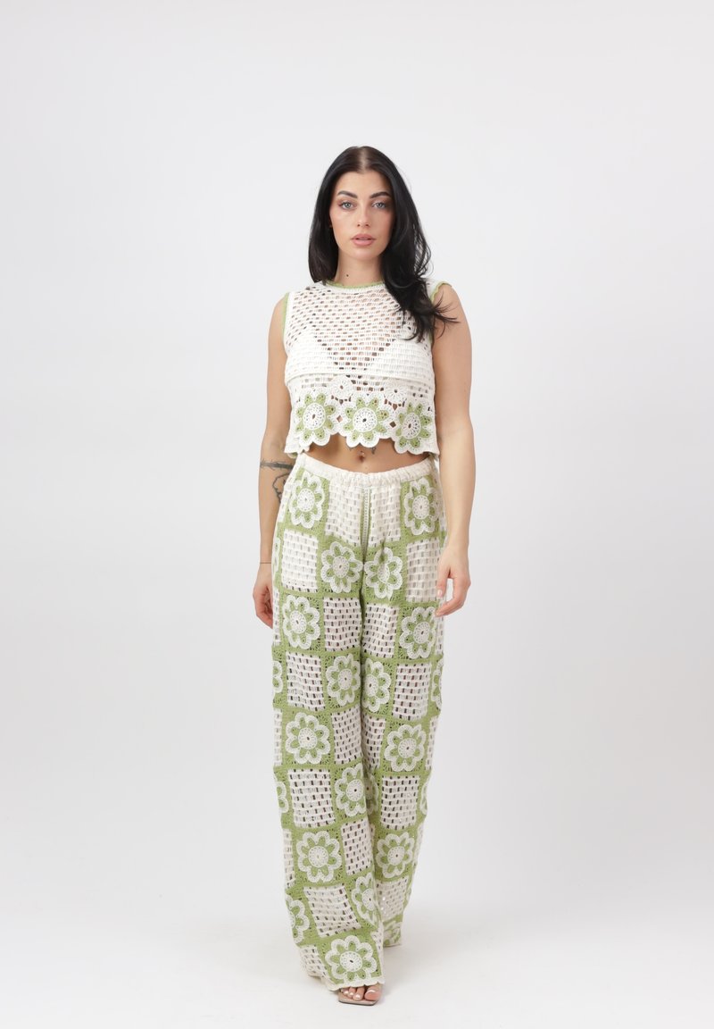 Vrouw draagt een witte en groene gehaakte crop top en broek met bloemmotieven, staand tegen een effen witte achtergrond.