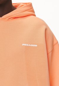 Oranžová mikina s kapucí z měkké látky s bílým logem "PEGADOR" na přední straně. Součástí je kapuce se šňůrkou a žebrované manžety.