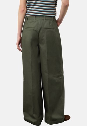 Persona vistiendo pantalones de pierna ancha color verde oliva con bolsillos de ojal en la parte trasera y un bolsillo con solapa abotonada al lado, combinado con una camisa a rayas.