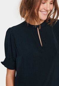 Blusa blu navy con scollatura a chiave e maniche corte a palloncino, caratterizzata da dettagli smock ai polsini. Tessuto dalla superficie liscia.