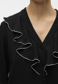 Sort bluse med V-udskæring, diskrete knapdetaljer og bølget kant i hvid, lavet af letvægtsstof med en struktureret overflade.