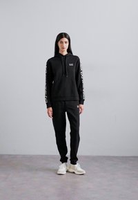 Ensemble de sweat-shirt à capuche et de pantalons de jogging noir, fabriqué à partir d'un tissu doux. La capuche est dotée de cordons de serrage et d'un logo ; le pantalon possède un écusson avec logo. Le modèle porte des baskets blanches.
