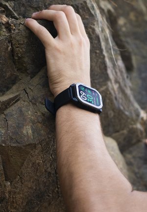 Hånd med smartwatch, der griber om en ru klippeoverflade og viser fysisk aktivitetsregistrering under udendørs klatring på klipper.