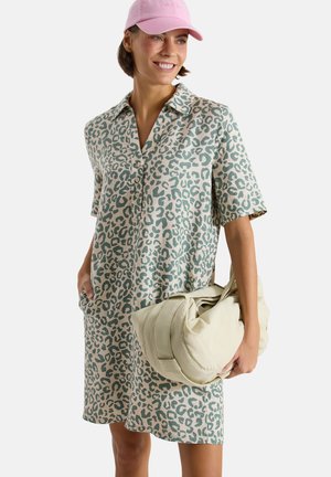 Femme portant une robe à manches courtes imprimée léopard verte et beige et une casquette rose, tenant un grand sac matelassé blanc cassé, souriante et regardant de côté.