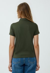 Polo in verde oliva con maniche corte e colletto, realizzato in un tessuto liscio. La vista posteriore mostra una silhouette aderente e nessun motivo visibile.
