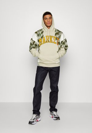 Beige hættejakke med "MARKET" i gul, prydet med camouflagemønstrede ærmer og hvide detaljer, parret med mørk denim jeans og sneakers.