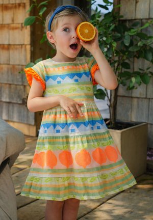 Young girl in colorful dress holds half an orange to her eye, standing outdoors near a plant with surprised expression.
