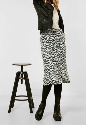 Jupe midi à motif noir et blanc en tissu fluide, associée à une veste en cuir noire ajustée et des bottines. Un tabouret en arrière-plan.