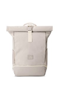 Mochila beige con tapa enrollable, correa y cierre con hebilla en color crema. Cuenta con un parche con logo y una textura suave, adecuada para uso casual.