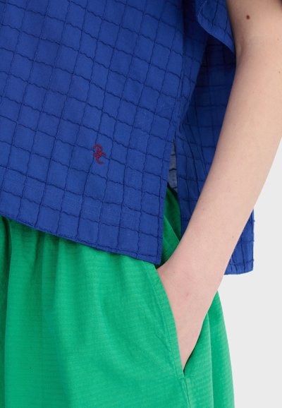 Gros plan sur une personne portant un haut bleu texturé avec une broderie rouge et un pantalon vert, la main dans la poche, sur un fond uni.