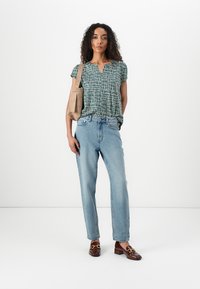 Groene blouse met een patroon en korte mouwen, lichtblauwe high-waisted jeans en bruine instappers. Draagt een beige tote bag.