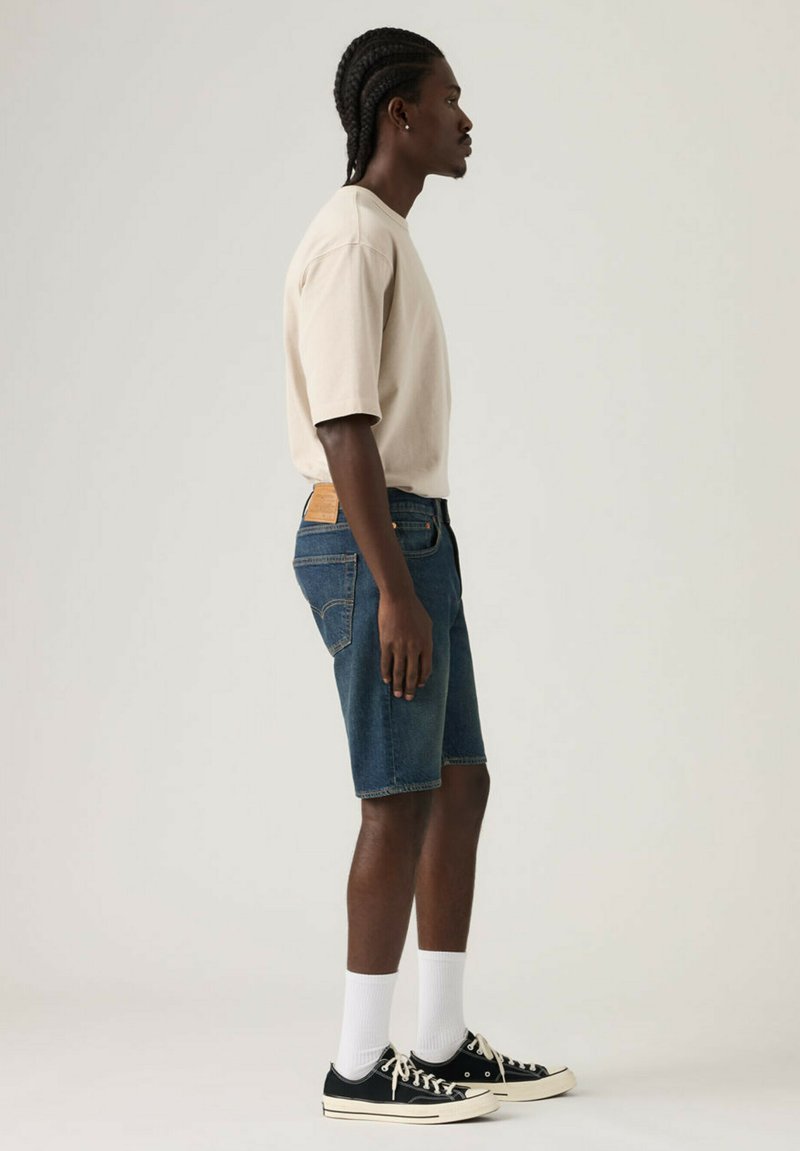 Homme aux cheveux tressés portant un t-shirt beige, un short en denim bleu, des chaussettes blanches et des baskets noires basses, debout de profil.