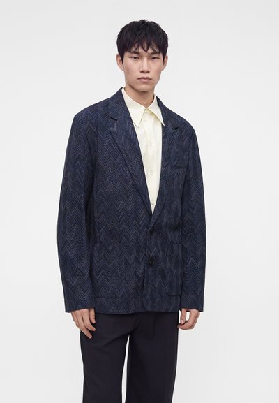 Jeune homme portant un blazer bleu marine à motif chevrons sur une chemise jaune pâle boutonnée et un pantalon noir, debout devant un fond uni.