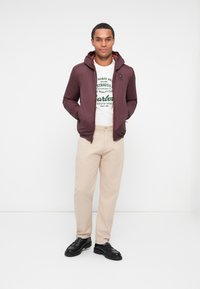 Chaqueta ligera color marrón burdeos con capucha, combinada con una camiseta gráfica blanca, pantalones crema y mocasines negros sobre un fondo sencillo.