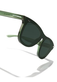 Vihreät muoviset aurinkolasit tummilla linsseillä, paksut neliönmuotoiset kehykset ja kiiltävä pinta. "HAWKERS"-logo hopeanvärisenä sangan kohdalla.