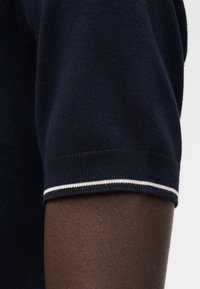 Μπλε ναυτικό πλεκτό ένδυμα με κοντά μανίκια και υφή, με λευκή τρέσα στο τελείωμα, εμφάνιση κοντινής λήψης του μανικιού.