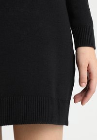 Zwarte gebreide jurk met een geribde onderrand en manchetten, met een close-up van de textuur en pasvorm rond de taille en hand.