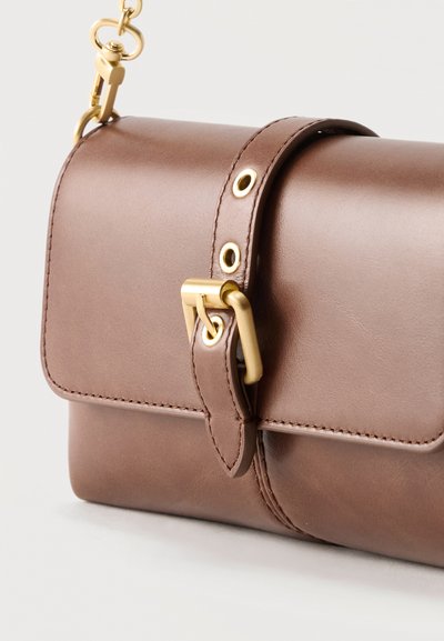 Καφέ δερμάτινη τσάντα χιαστή με χρυσή αγκράφα και λεπτομέρειες αλυσίδας στο λουρί, εμφανίζεται από κοντινή πλαϊνή γωνία.